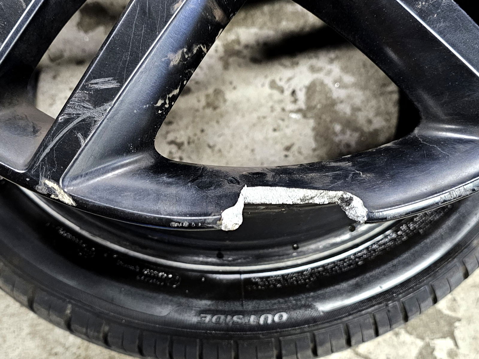 Scratched black car rim with significant curb damage, showing exposed silver metal underneath the dark finish.